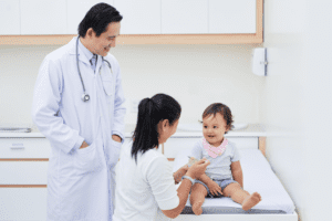 A doctor watches as a mother feeds a baby sitting on an exam table during an oral food challenge to test for food allergy